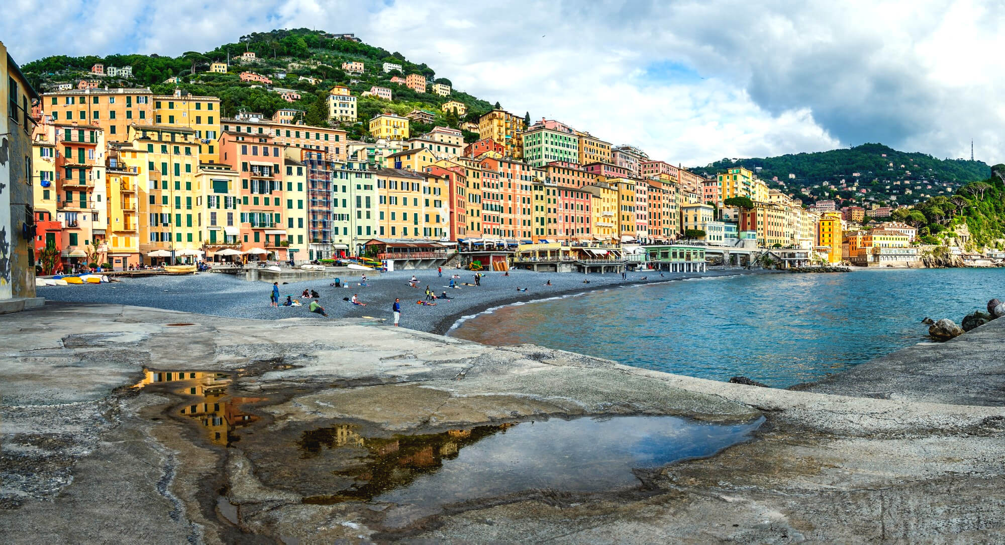 Colorful harbor town with pastel buildings along a pebble beach and blue sea, hillside in the background.