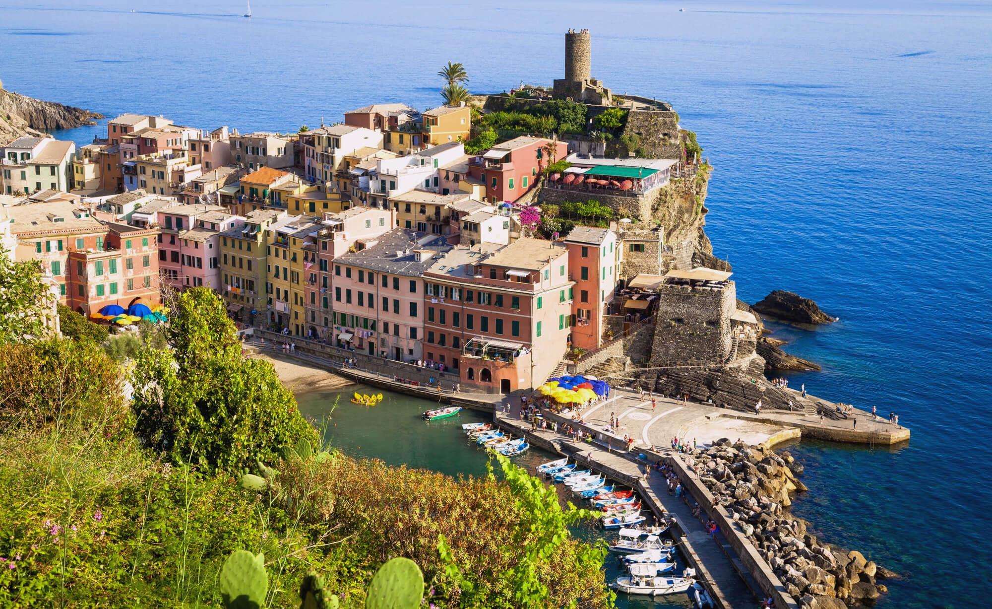Colorful coastal town perched on a cliff, with pastel buildings, a stone tower on the peninsula, and a busy harbor with boats and umbrellas by the water.