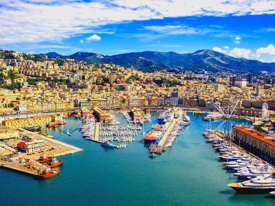 Panoramic view of a coastal city with a busy harbor, colorful hillside buildings, and distant mountains under a blue sky.