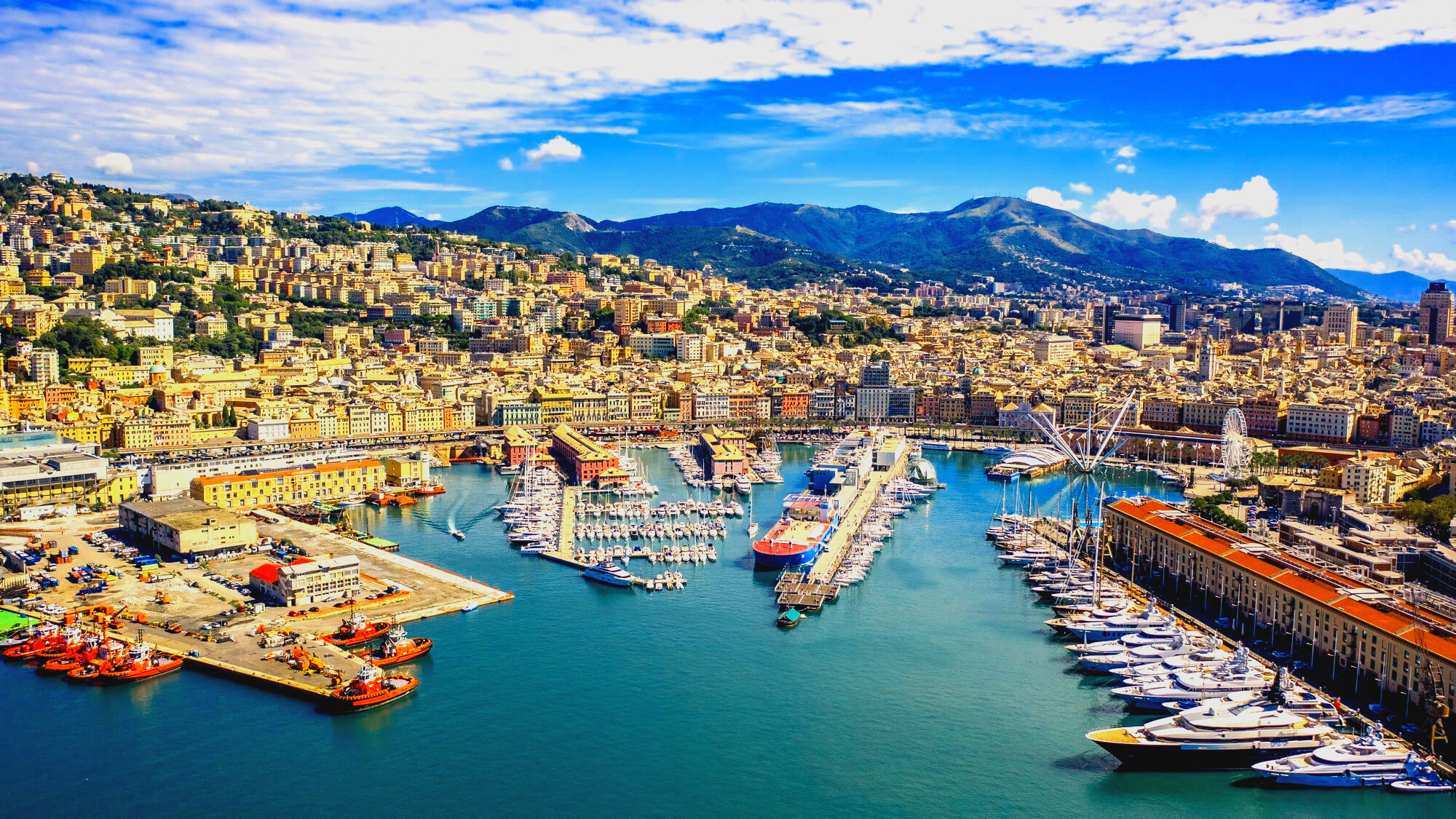 Panoramic view of a coastal city with a busy harbor, colorful hillside buildings, and distant mountains under a blue sky.