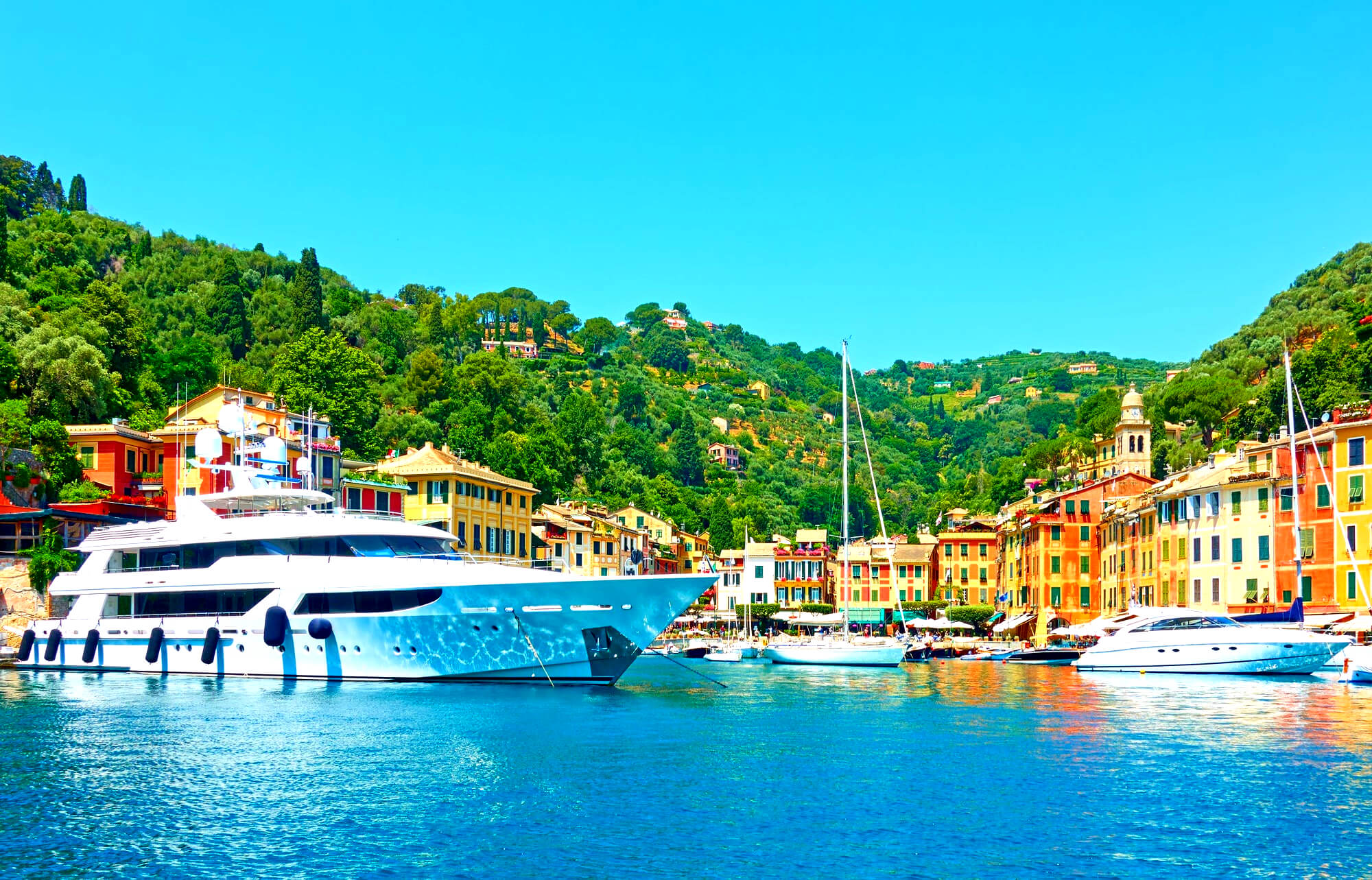 Harbor scene with a large white yacht, colorful buildings along the waterfront, and green hills in the background under a bright blue sky.