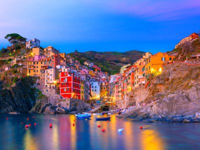 Colorful coastal hillside town at dusk with yellow and red buildings along rocky shore and calm water reflection