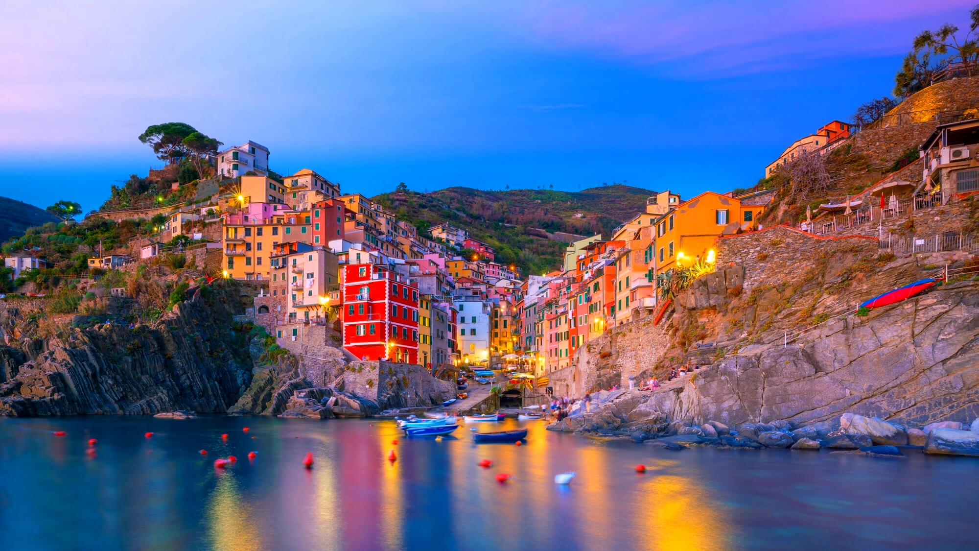 Colorful coastal hillside town at dusk with yellow and red buildings along rocky shore and calm water reflection